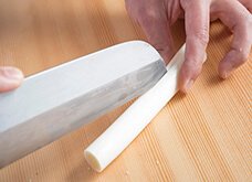 約10cmに切り落とした長ねぎを親指と人差し指でつまんで押さえ、包丁の切っ先で縦半分に切る。