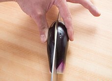 親指と人差し指でなすの丸みのあるほうをつまむようにして押さえ、包丁の切っ先で縦半分に切る。