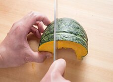 かぼちゃの丸みのあるほうを奥にしてまな板に置き、かぼちゃを持つ指で押さえ固定しながら2/3～半分ほどに切り落とす。